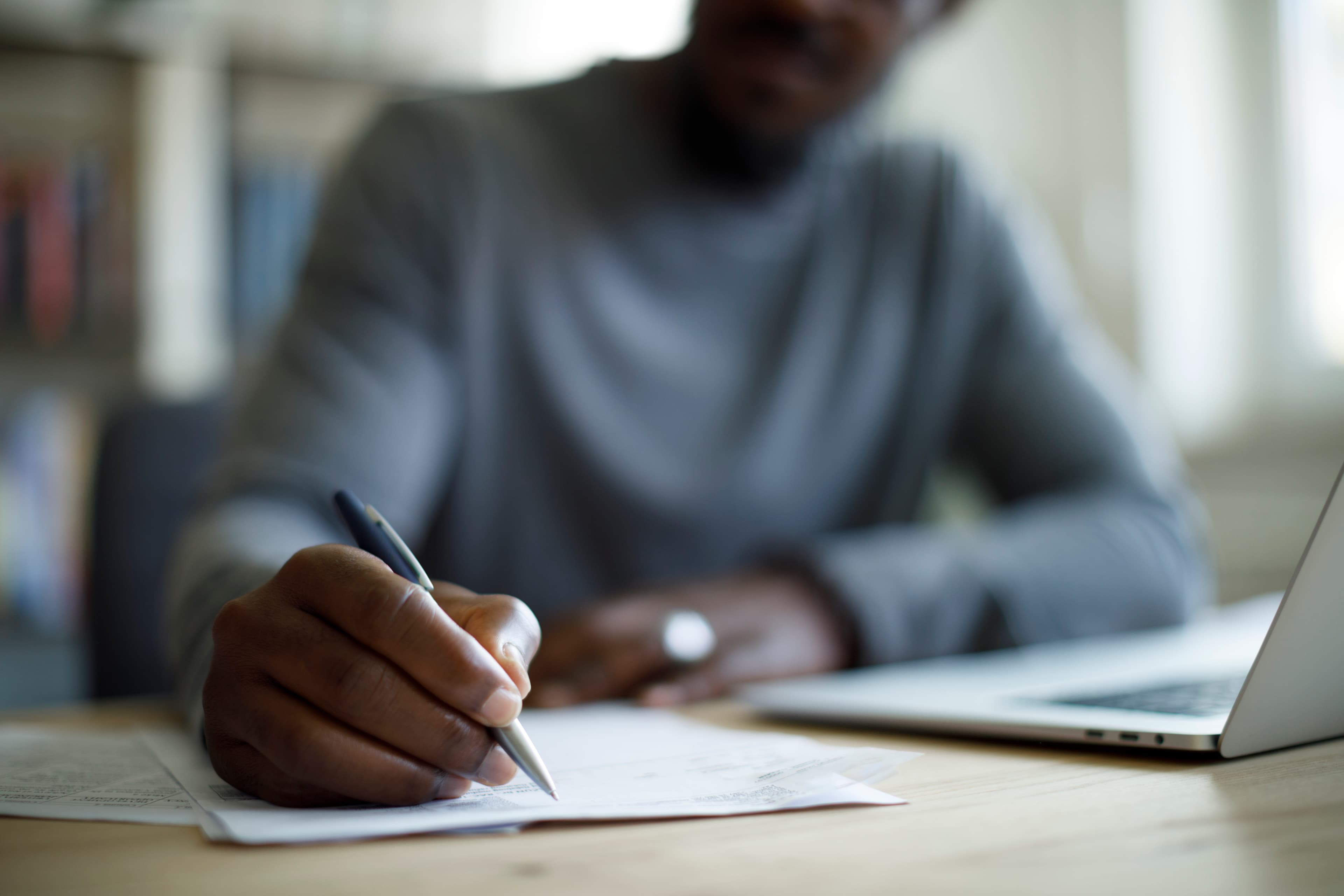 A black person writing with their right hand uses a pen to write on a piece of paper set next to an open laptop. The image is blurred except for the person's writing hand.